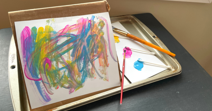 A piece of cardboard is folded in half and taped to a tray to mimick an easel. Paint brushes and cotton swabs are used to paint a paper taped to the cardboard.