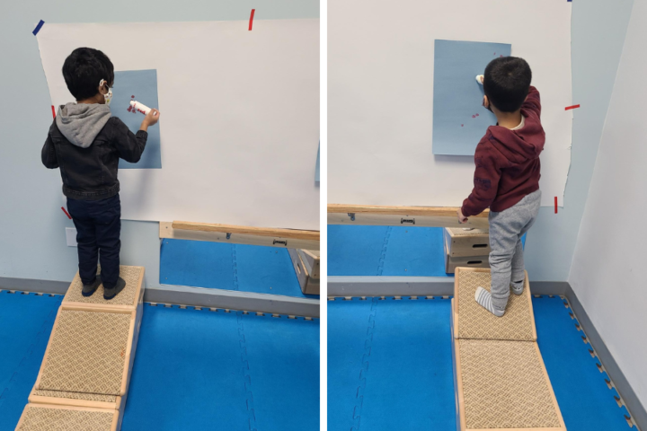Two photos of a preschool aged child standing on a step stool while colouring on paper taped to the wall in front of them.