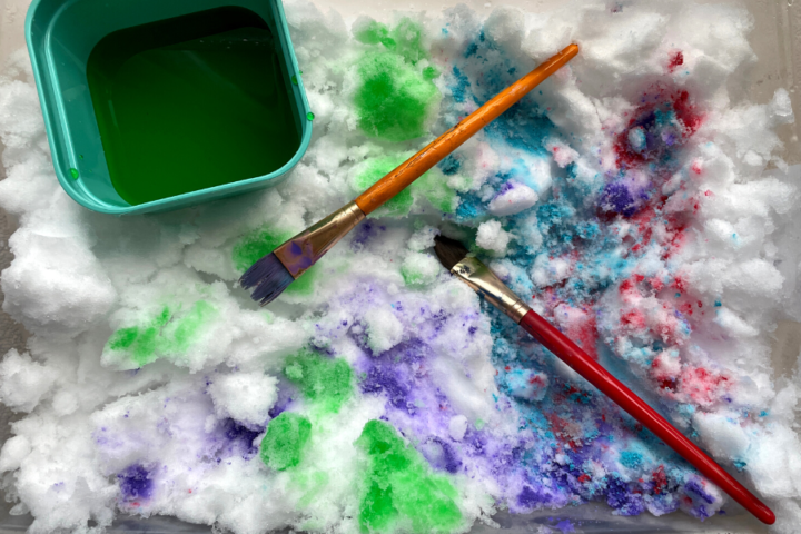 Colourful snow inside a bin with paint brushes and a bowl filled with water and food colouring.